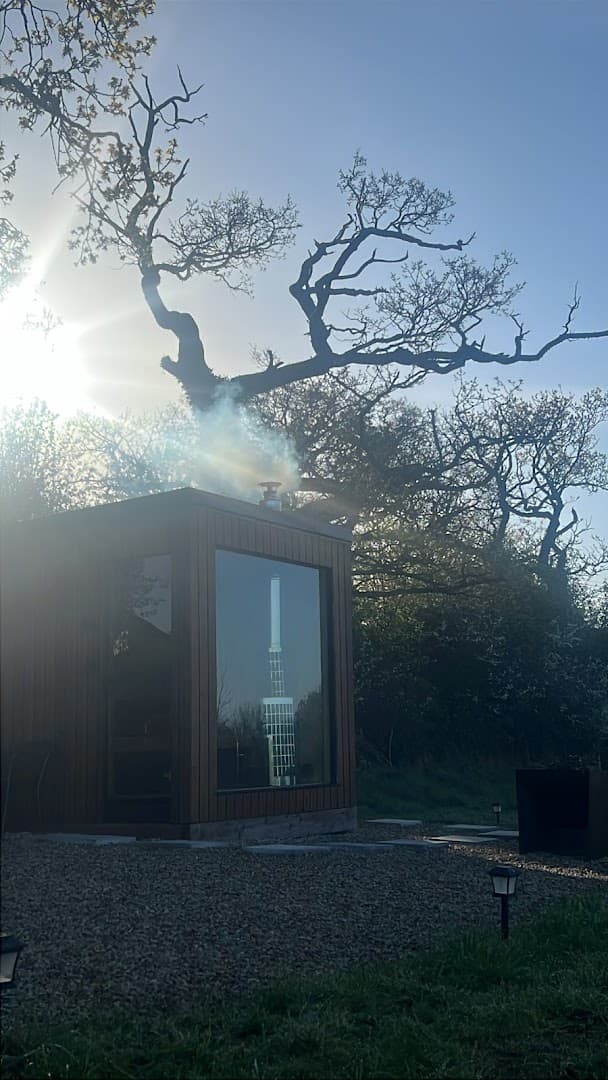 A wooden sauna with smoke rising from its chimney sits on a gravel path outdoors beneath large bare trees.