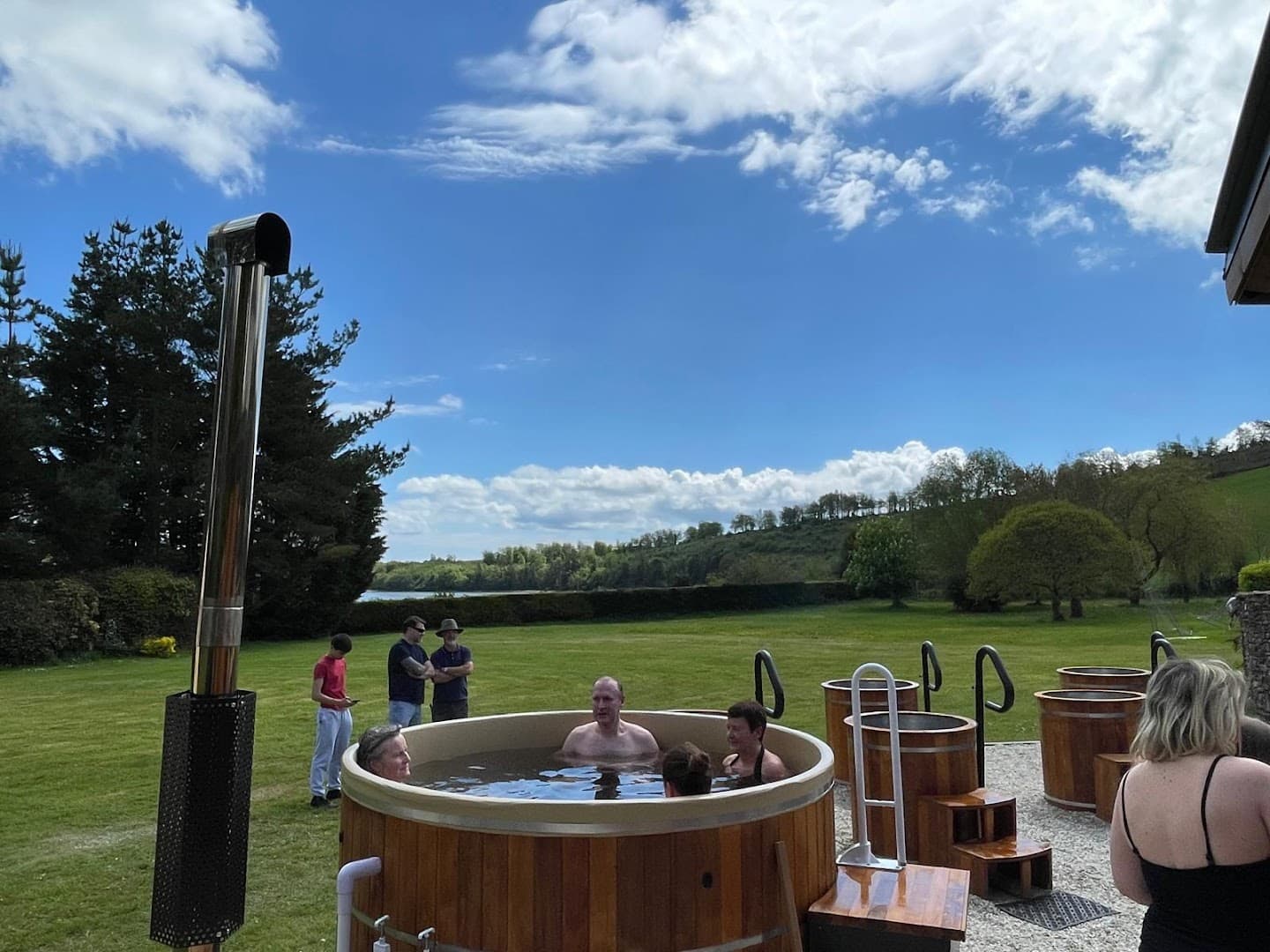 Several people relax in a wooden hot tub outdoors on a sunny day, with a landscape view and other small tubs nearby.