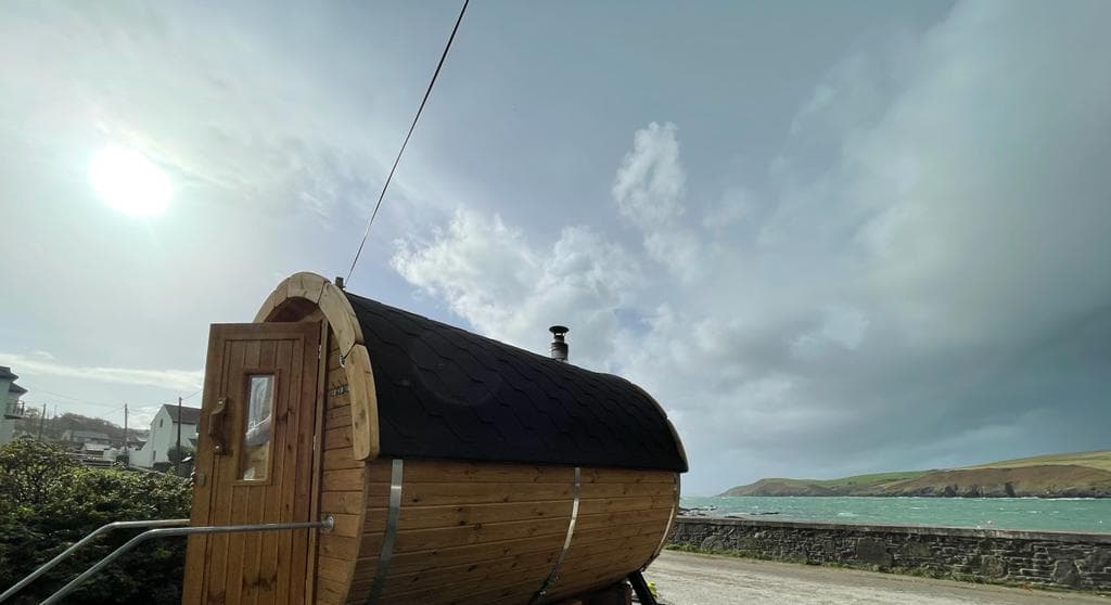 A wooden barrel sauna sits near the coast with a partial view of the sea and sky.