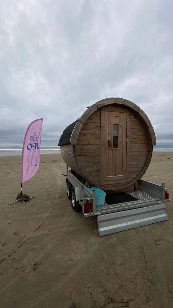 A wooden barrel sauna on a flatbed trailer sits on a sandy beach next to the sea under an overcast sky.