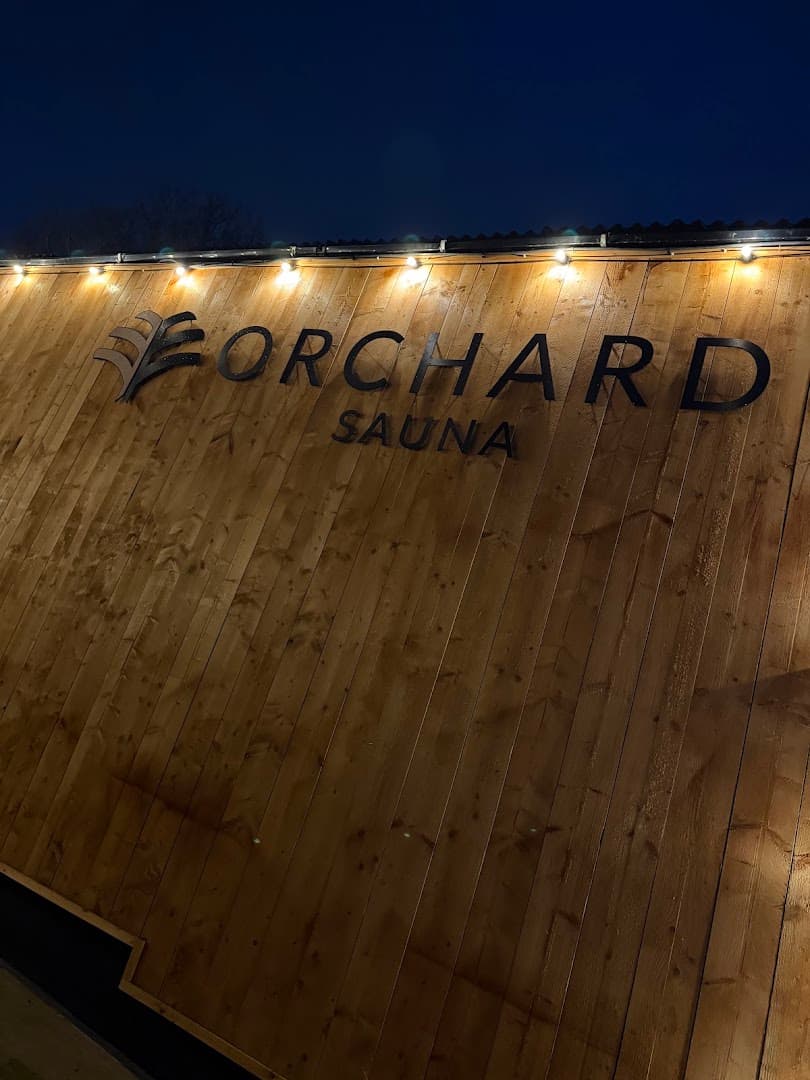 The wooden exterior wall of Orchard Sauna features its name in large black letters and is illuminated by string lights.
