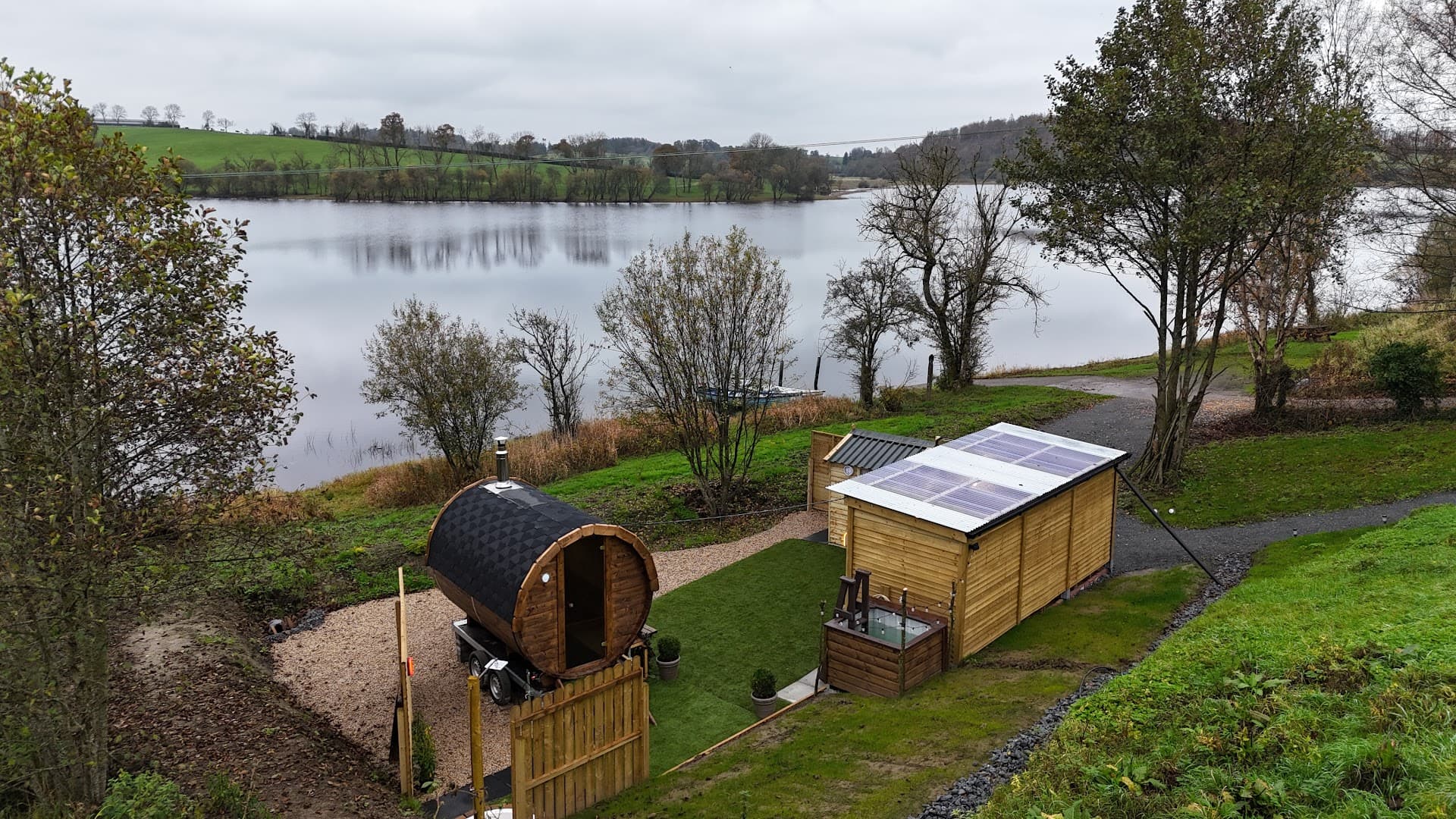 An elevated view of a wooden barrel sauna and a shed with solar panels overlooking a calm lake in Killashandra.