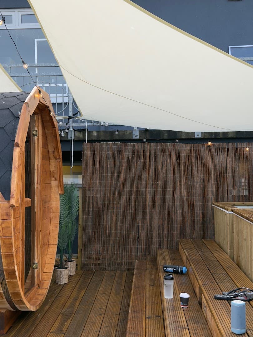 An outdoor wooden barrel sauna stands on a deck with steps beneath a beige shade sail.