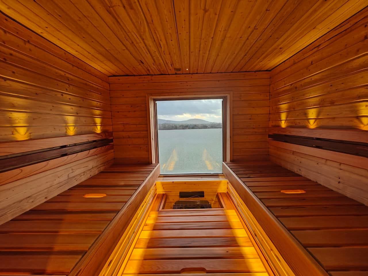 Interior view of a wooden sauna featuring slatted benches, warm lighting, and a square window looking out over water.
