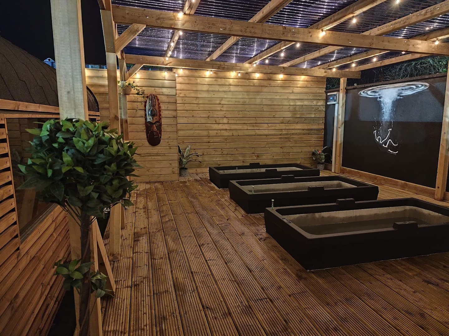 Outdoor wooden deck with ice baths and a roof under string lights at night.