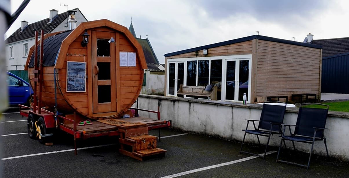A wooden barrel sauna on a trailer sits parked outside a modern building with glass doors in Kilmovee.