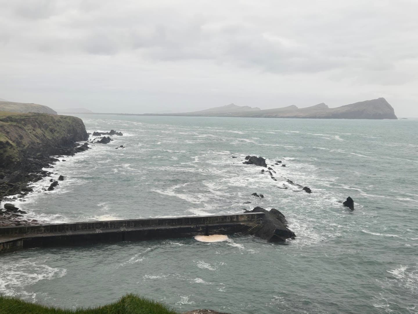 An elevated view of a concrete pier extending into the rough sea on an overcast day in Ireland.