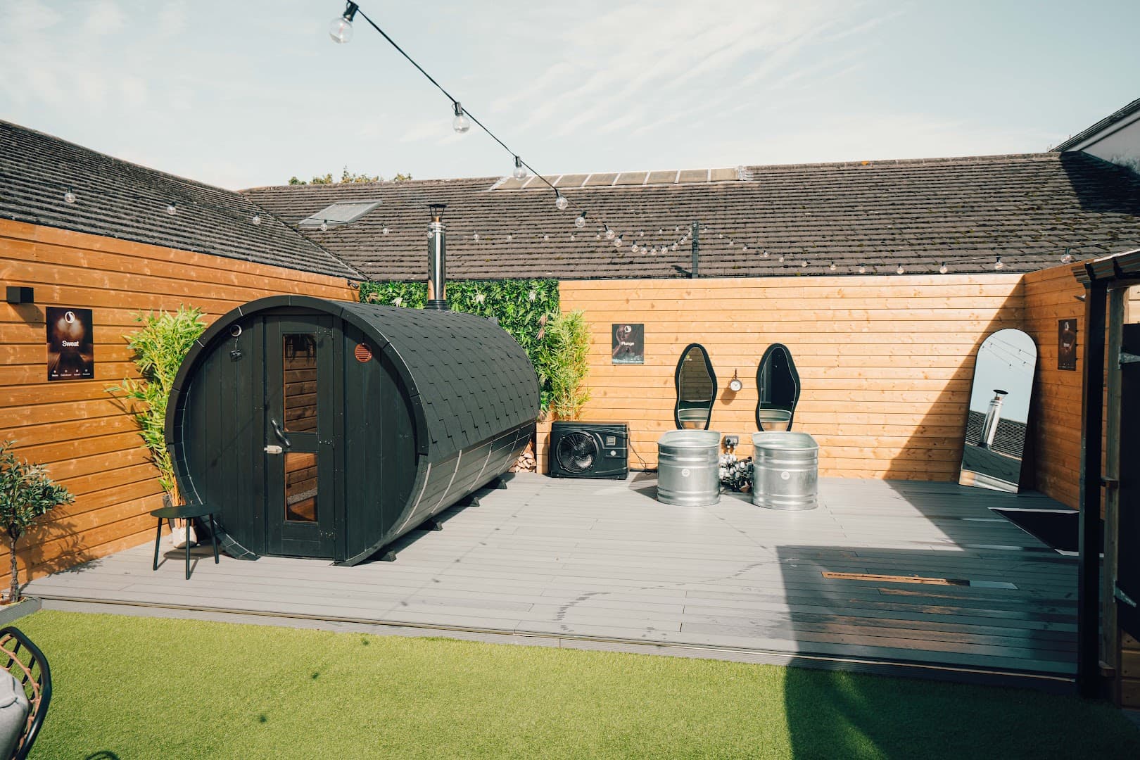 A barrel sauna and cold plunge tubs sit on a wooden deck in an outdoor wellness space.