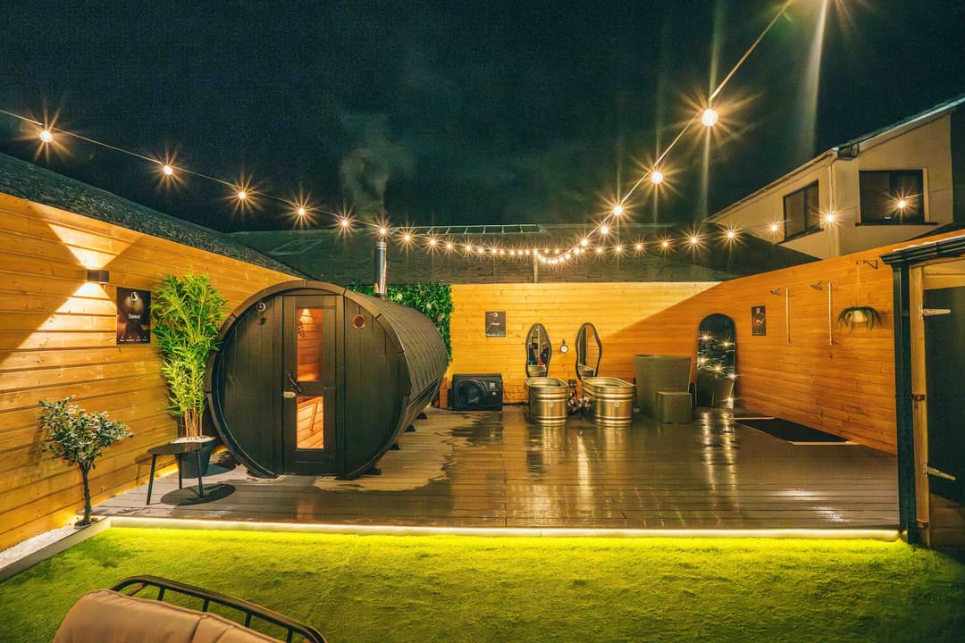 Night view of a barrel sauna on a wooden deck with string lights and two metal tubs.