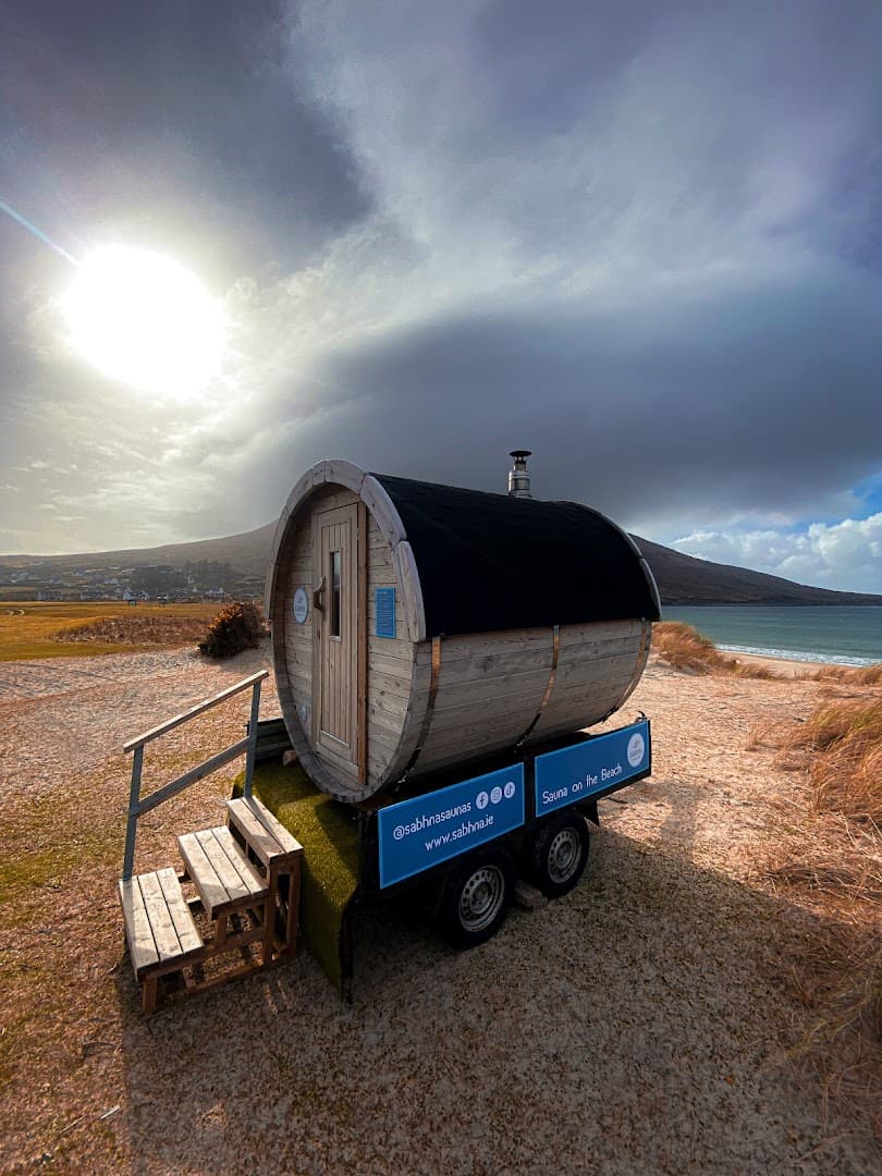 A mobile barrel sauna on a trailer sits on a sandy beach next to the ocean under a partly cloudy sky.