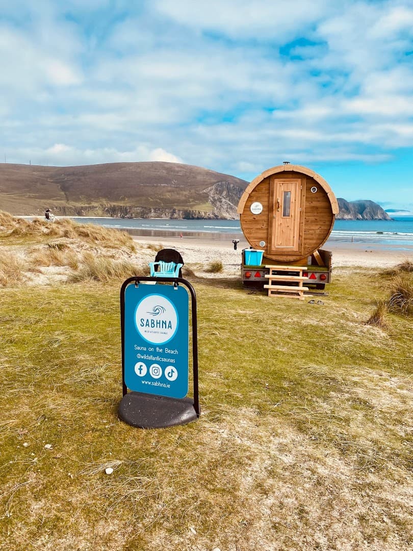 A barrel sauna on a trailer sits near grassy dunes and a sandy beach with the sea in the background.