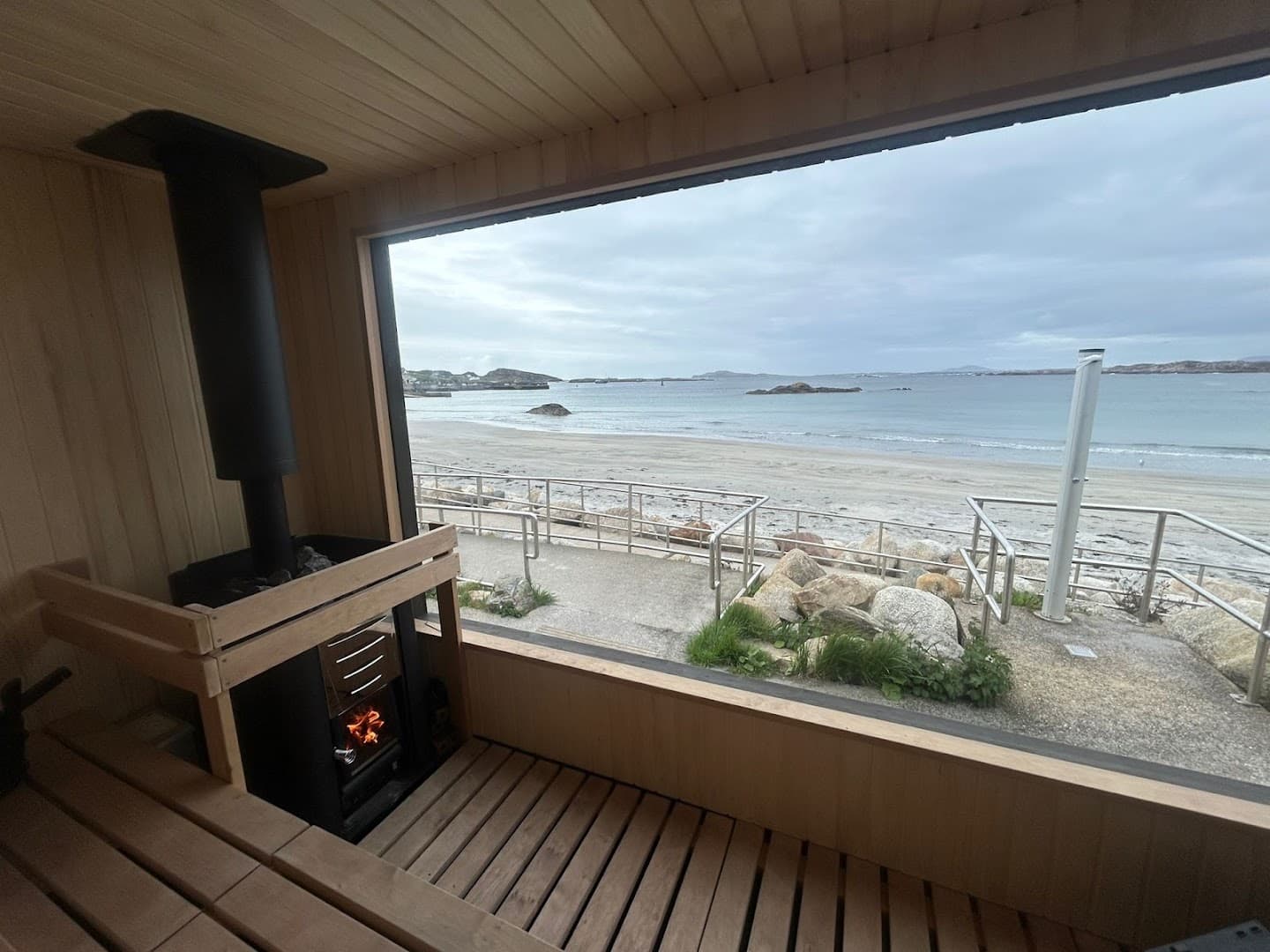 Wooden sauna interior with wood-burning stove looking out onto a sandy beach and sea.
