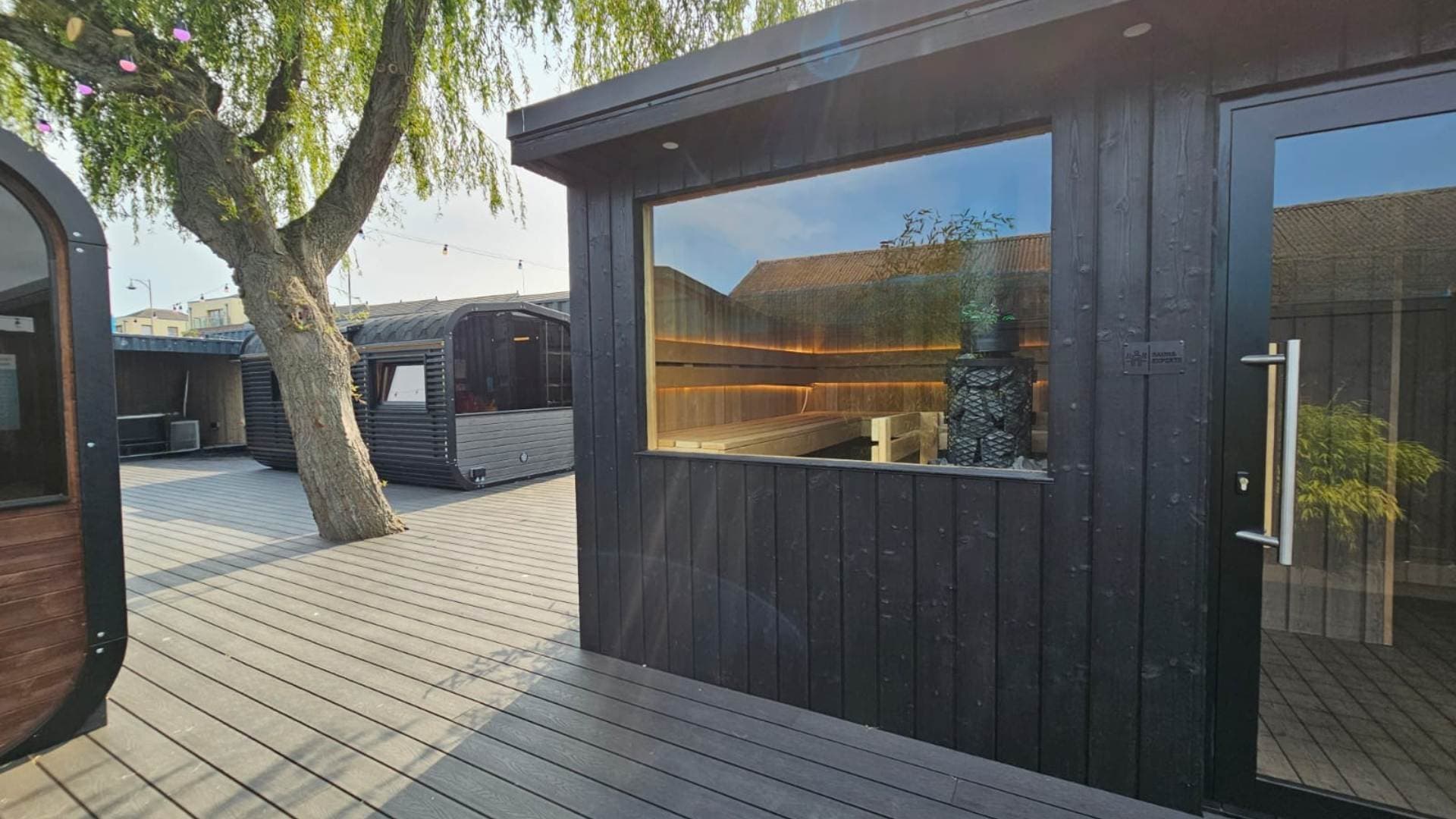 Exterior of a black wooden sauna building with a large glass window and door, on an outdoor wooden deck.