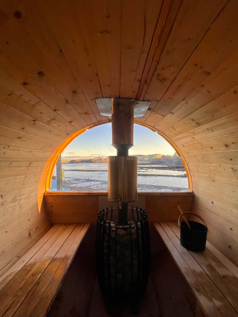 Interior view of a wooden barrel sauna with a stove, benches, and a large window facing the sea.