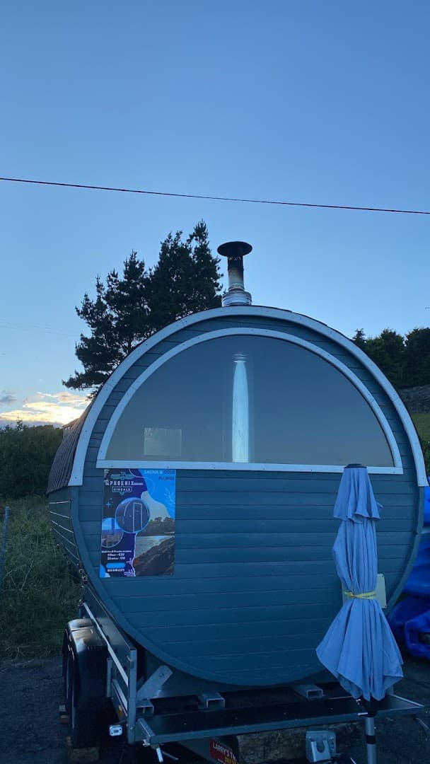 A wooden barrel-shaped sauna on a trailer sits outdoors with a closed umbrella beside it against a clear sky.