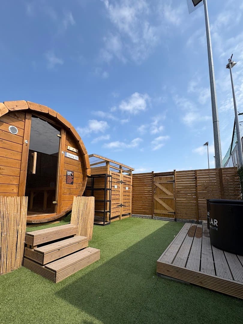 A barrel sauna stands outdoors next to a small wooden deck, storage shed, and fenced area under a partly cloudy sky.