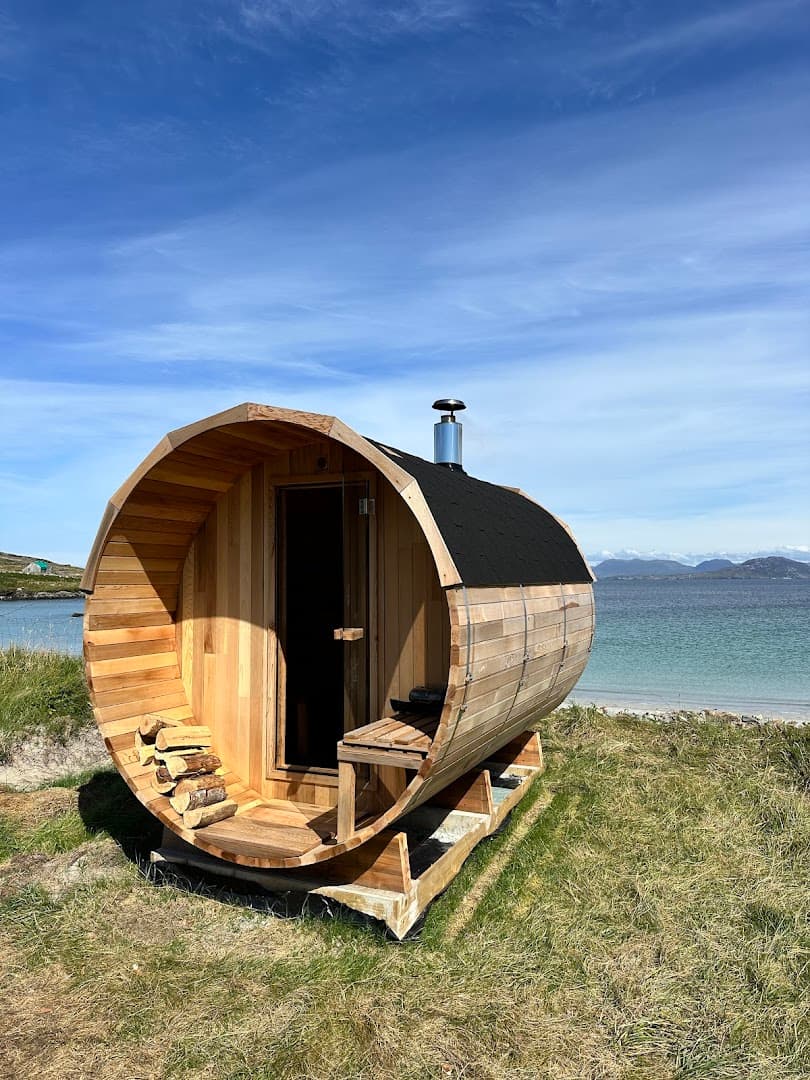 A wooden barrel sauna on a grassy coastline overlooks a blue sea and distant hills.