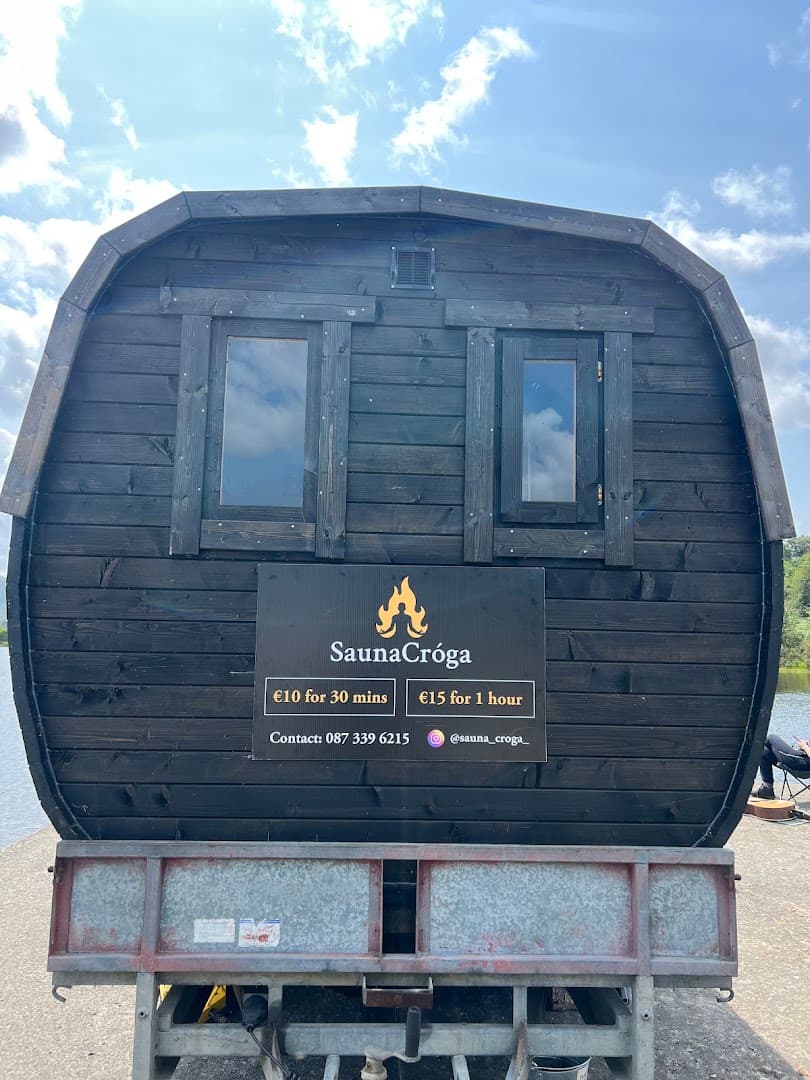 A black wooden barrel sauna on a trailer with price and contact signs stands near a body of water under a cloudy sky.