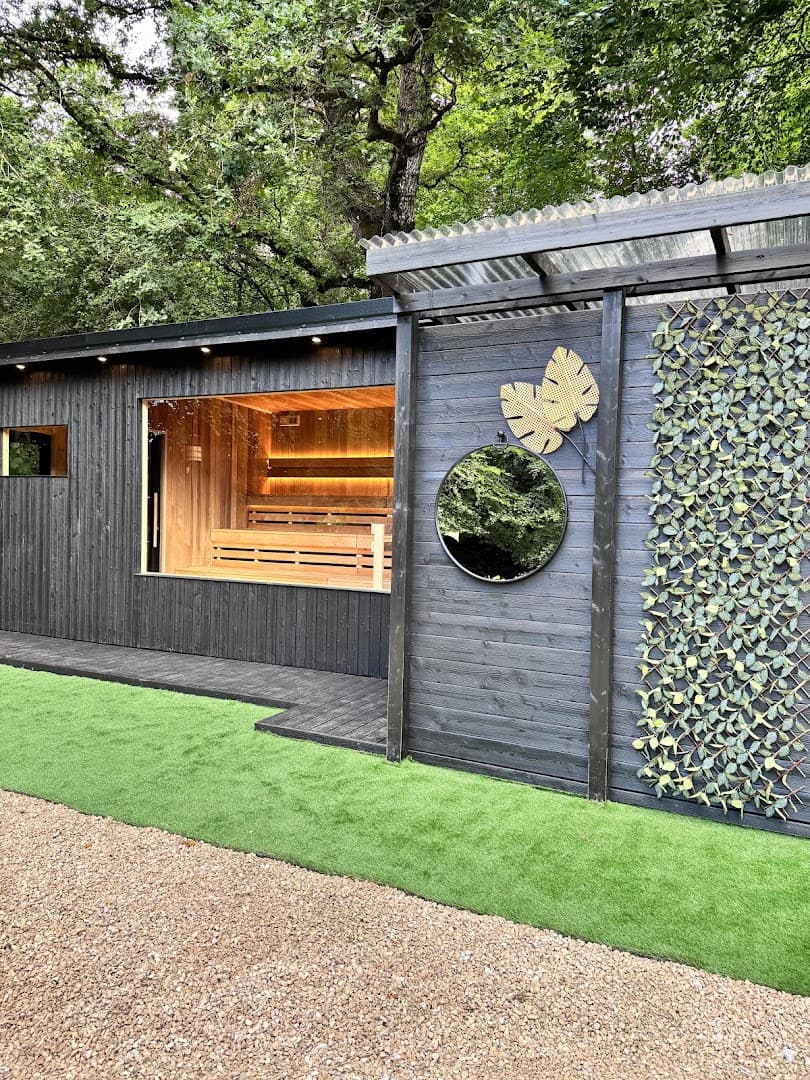 A black wooden sauna structure stands outdoors with a large glass window, a round mirror, and decorative leaves on its wall.