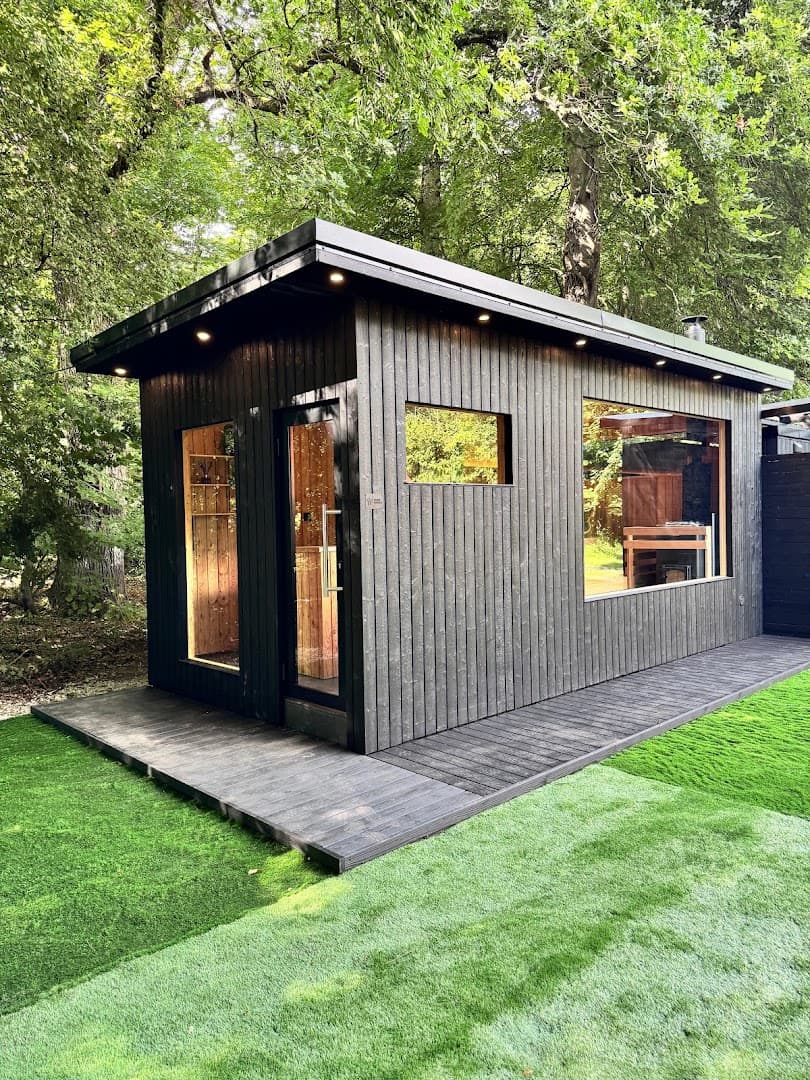 A black wooden sauna building stands on a deck amidst trees and green grass.