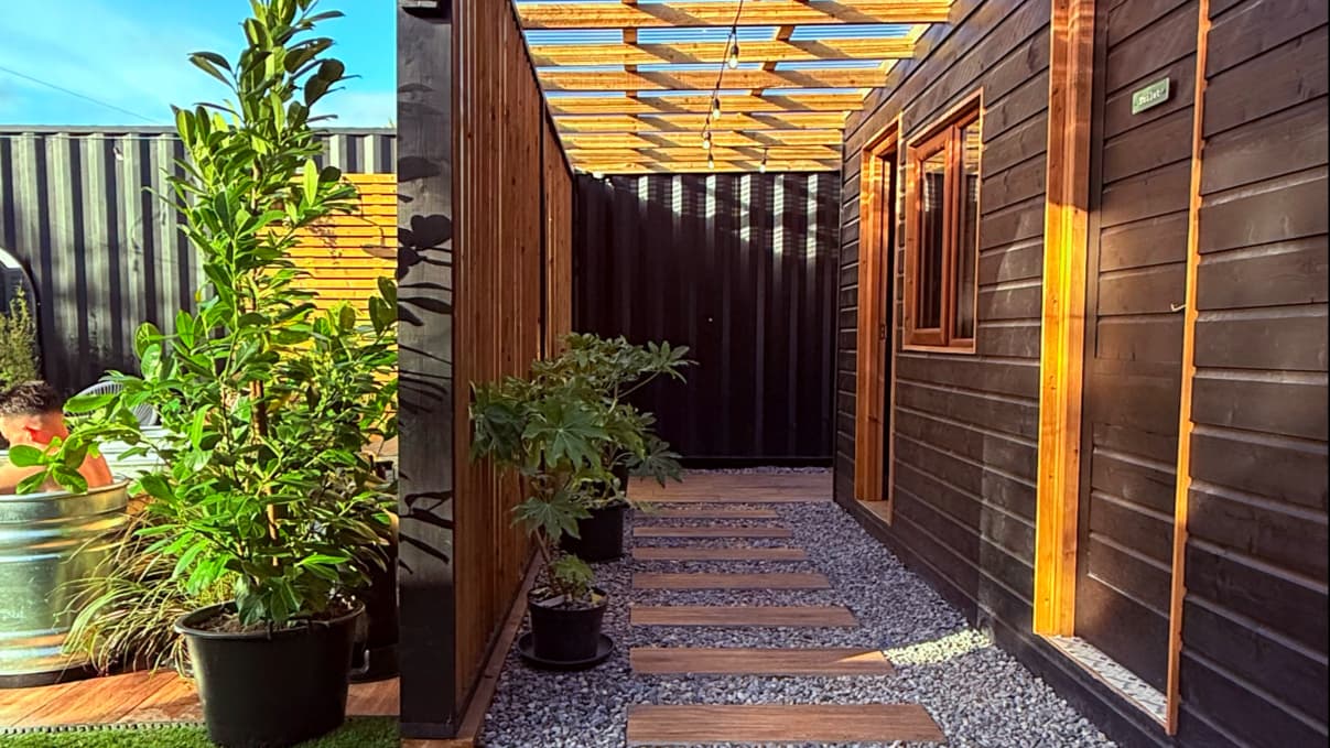 A narrow wooden path with stepping stones leads between a dark wooden wall and a structure with an open pergola.