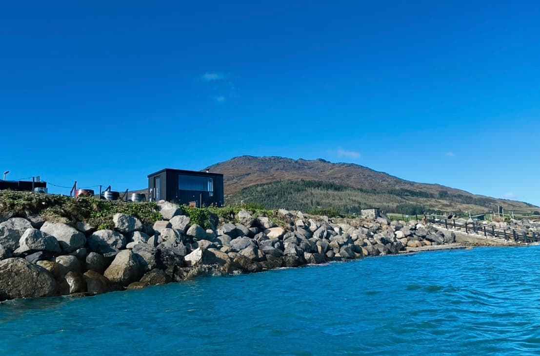 A dark sauna building sits on a rock-lined coast beside the sea, backed by a large mountain and a blue sky.