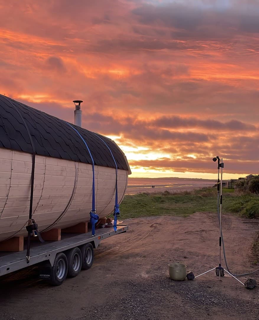A barrel sauna on a trailer sits by a beach at sunset with a shower stand nearby.