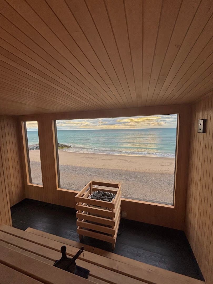 Inside a wood-paneled sauna, a large window frames a sandy beach and the ocean.