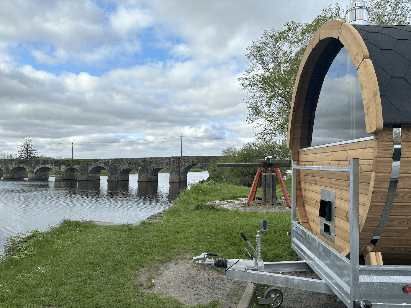 A barrel sauna on a trailer sits by a grassy riverbank with a stone bridge in the distance.