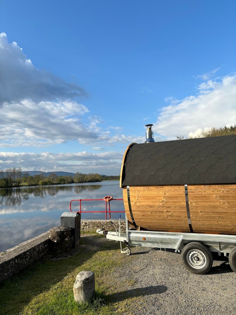 A wooden barrel sauna on a trailer sits by a reflective river under a blue sky.
