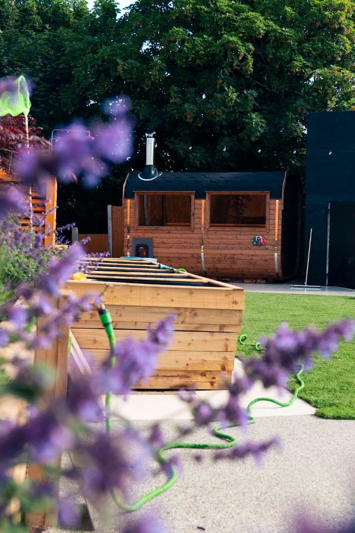 A wooden barrel sauna with a black roof sits in a grassy backyard, framed by purple flowers.