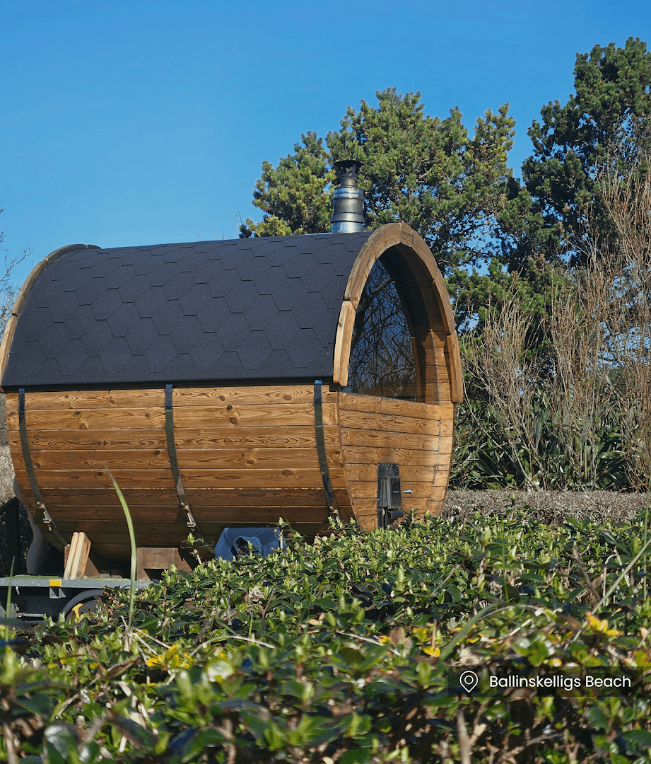 A wooden barrel-shaped sauna with a black shingle roof sits in front of green bushes under a blue sky.