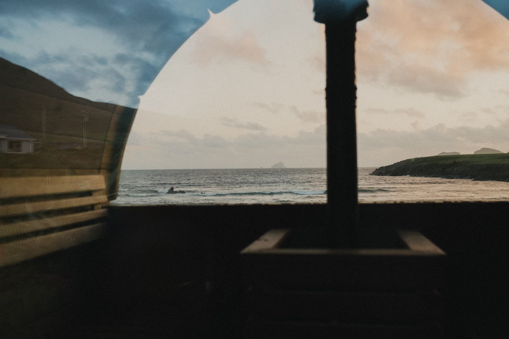 Inside a sauna with a dark interior, a large window looks out at a choppy sea and distant headlands under a cloudy sky.
