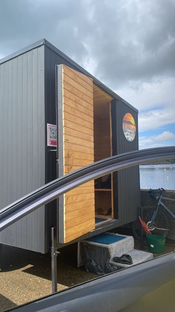 A small dark grey sauna structure with a wooden door stands near the sea under a cloudy sky.