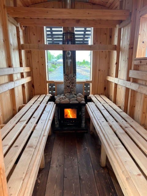 An interior view of a wooden sauna featuring slatted benches, a central wood-burning stove filled with stones, and a large window at the back.