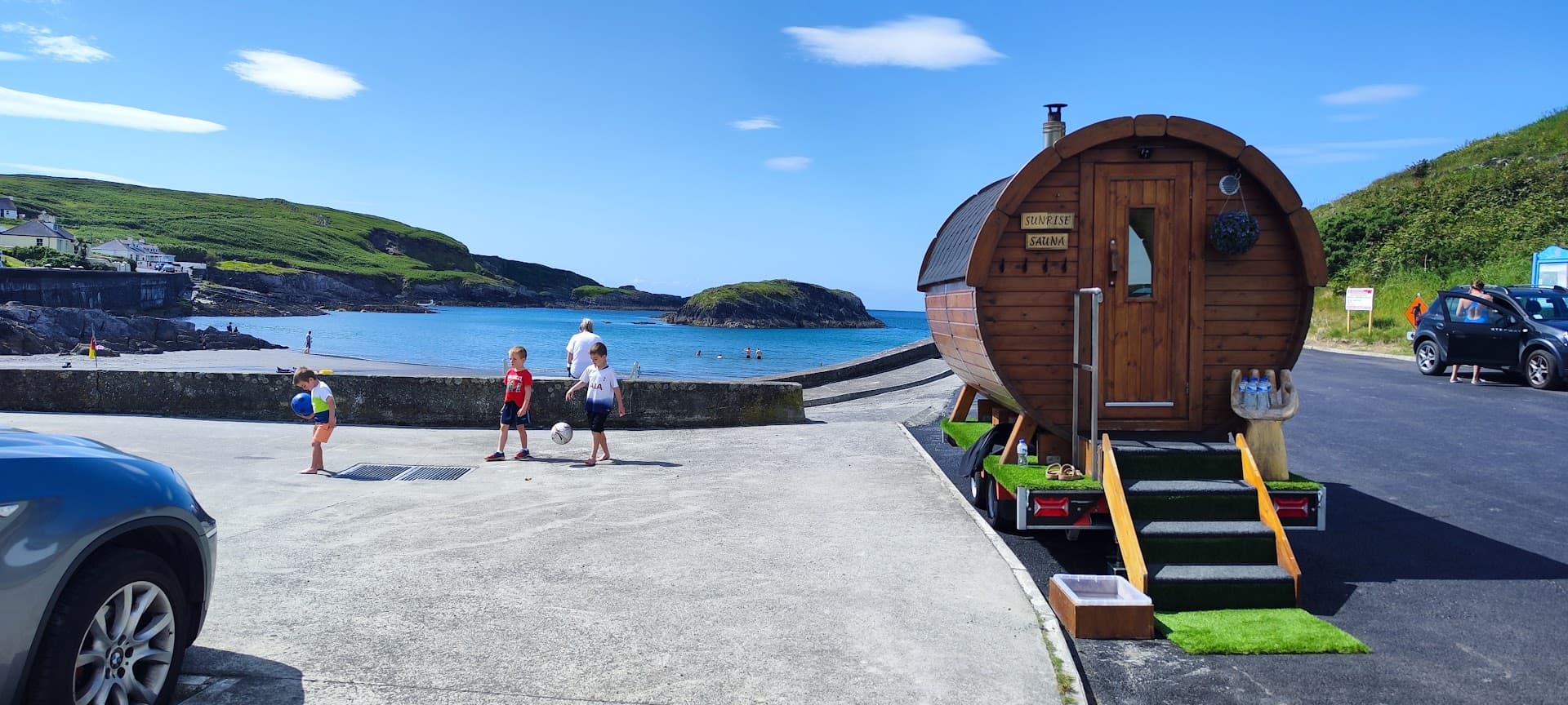 A barrel-shaped sauna on a trailer is parked near a beach where children play.