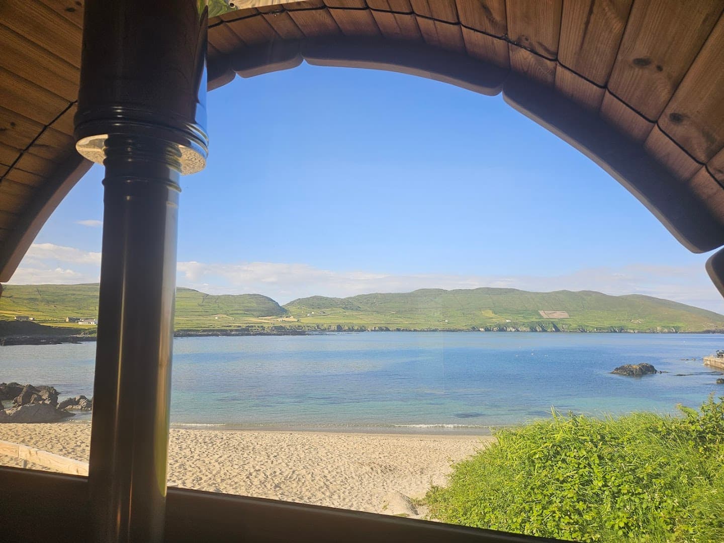 Looking out from inside a sauna towards a sandy beach, calm sea, and green hills under a blue sky.