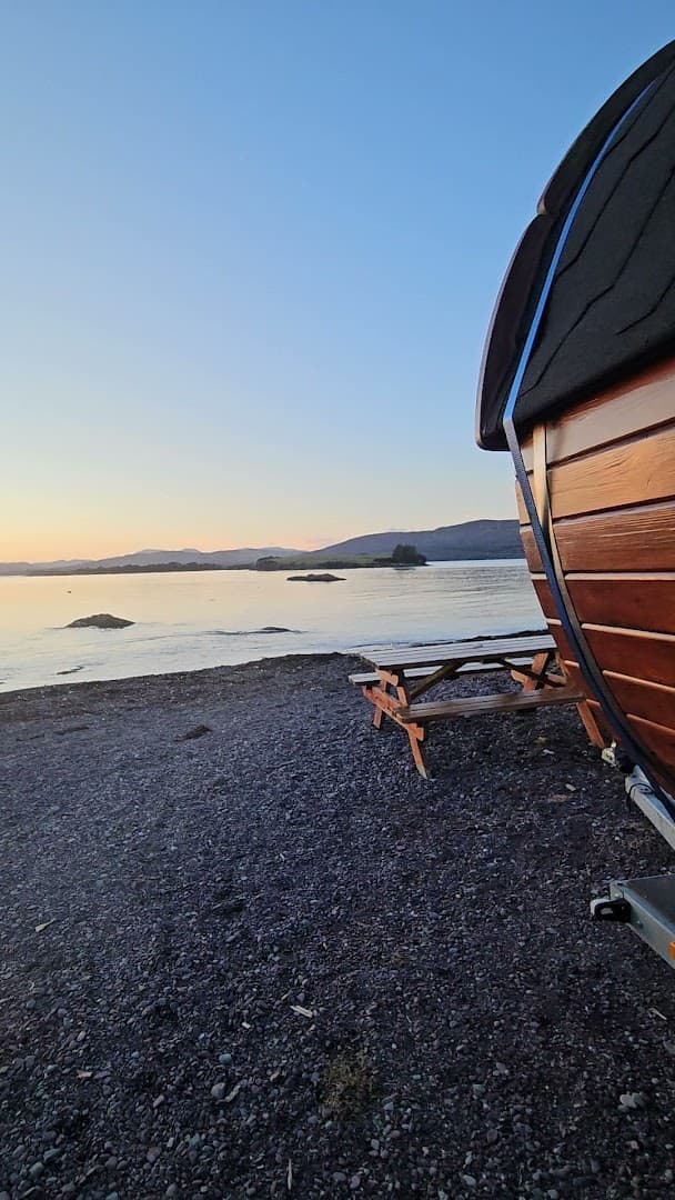 A wooden barrel sauna on a pebbled beach overlooks the sea during sunset.