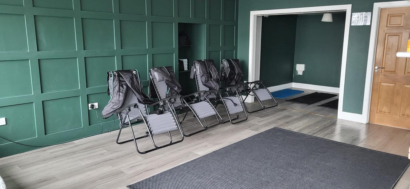 A room with a dark green panelled wall, a row of folded loungers, grey flooring, and a doorway.