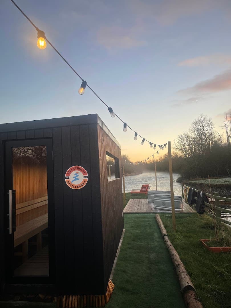 A black sauna structure with a glowing interior stands outdoors on grass next to a wooden deck with metal tubs, under string lights and a dusk sky.
