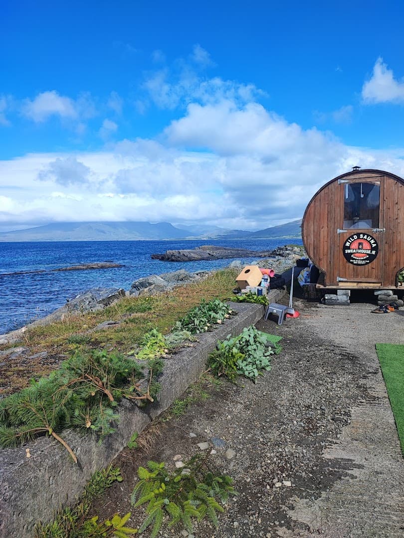 A wooden barrel sauna sits by the sea on a rocky coast under a cloudy blue sky.