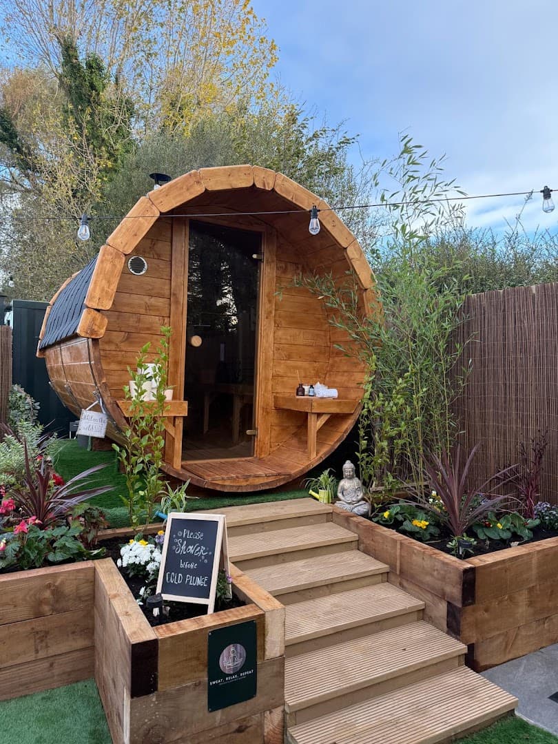 A wooden barrel sauna with a glass door stands in a garden with wooden steps, raised flowerbeds, and string lights.