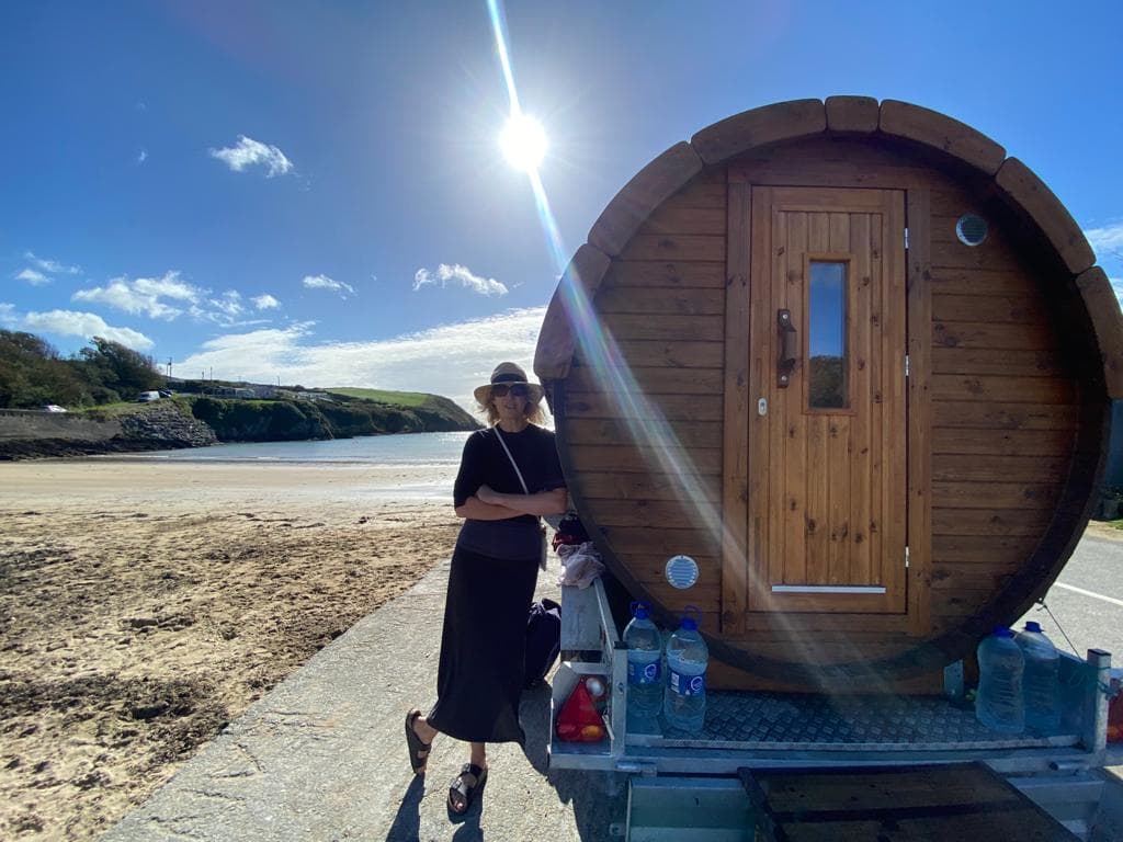 A woman stands beside a barrel sauna on a trailer parked near a sandy beach and ocean under a sunny sky.
