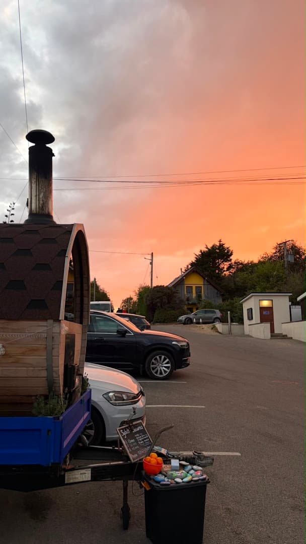 A barrel sauna on a trailer is parked in a paved lot beneath a vibrant orange and grey sunset sky.