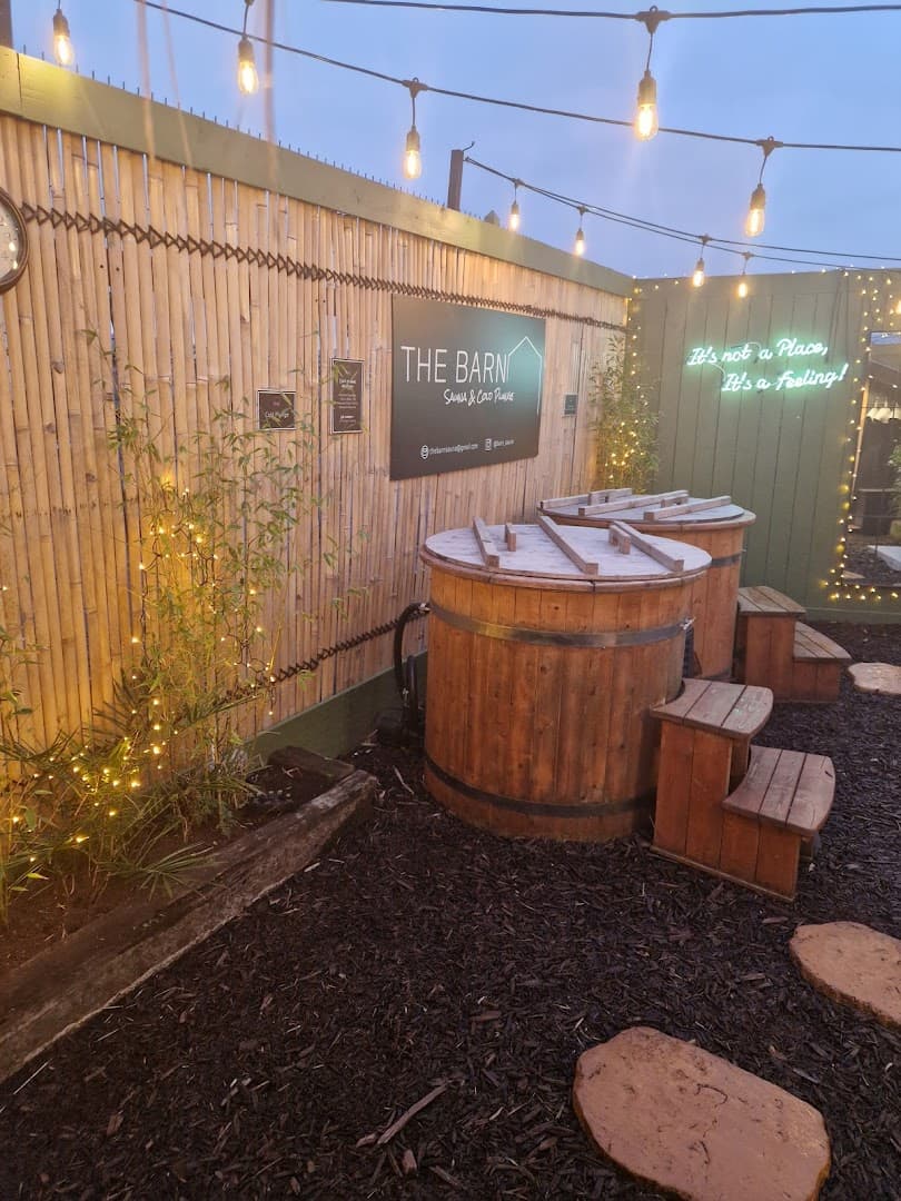 Two wooden cold plunge tubs sit on dark mulch next to a bamboo fence with "THE BARN" sign.