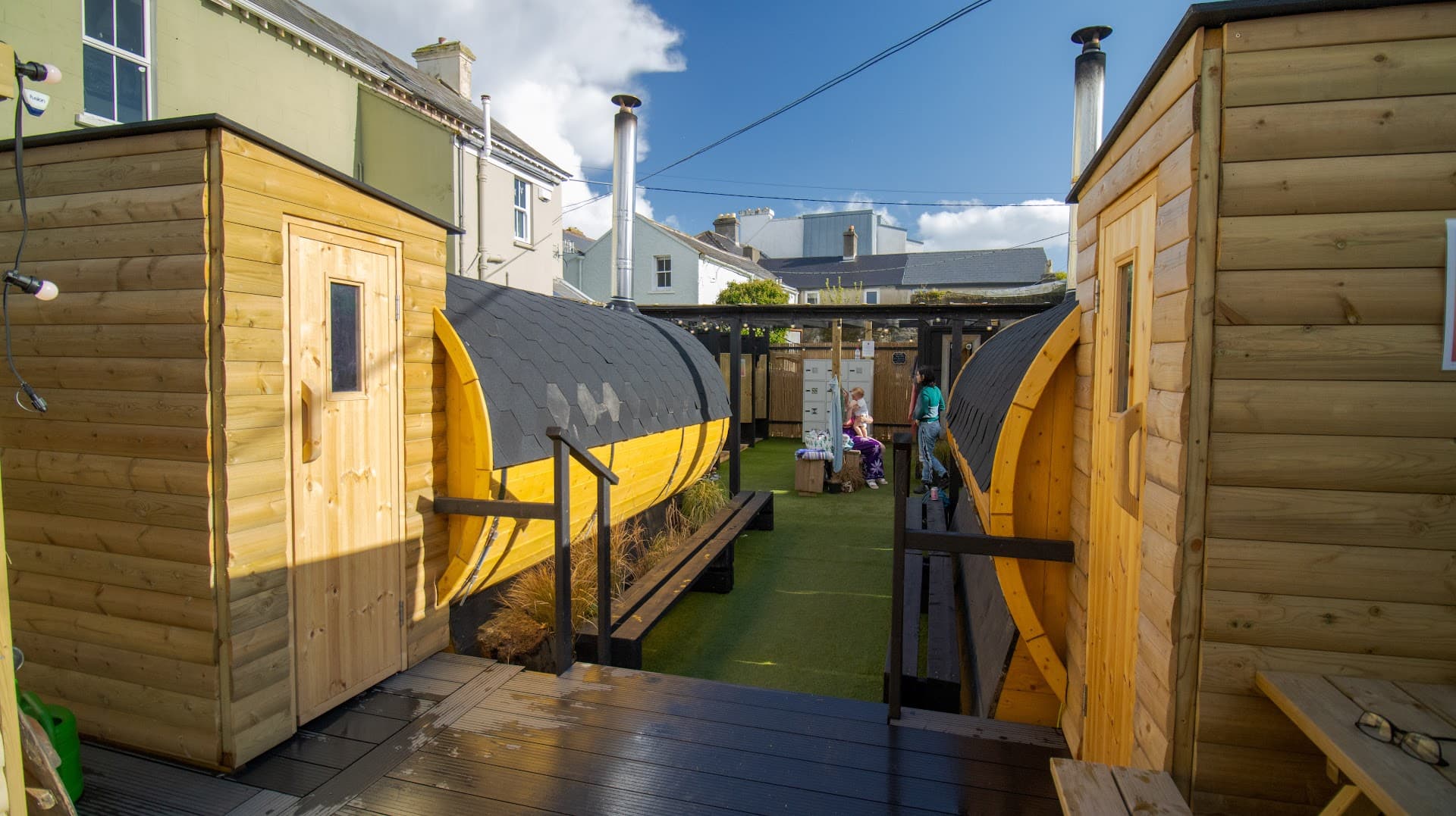 Two barrel saunas face each other on an outdoor deck with a grassy courtyard in between.