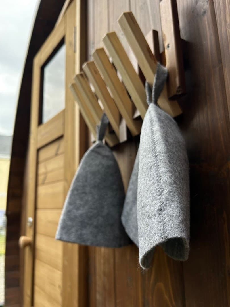 Two grey felt sauna hats hang on a wooden wall rack next to a sauna door.