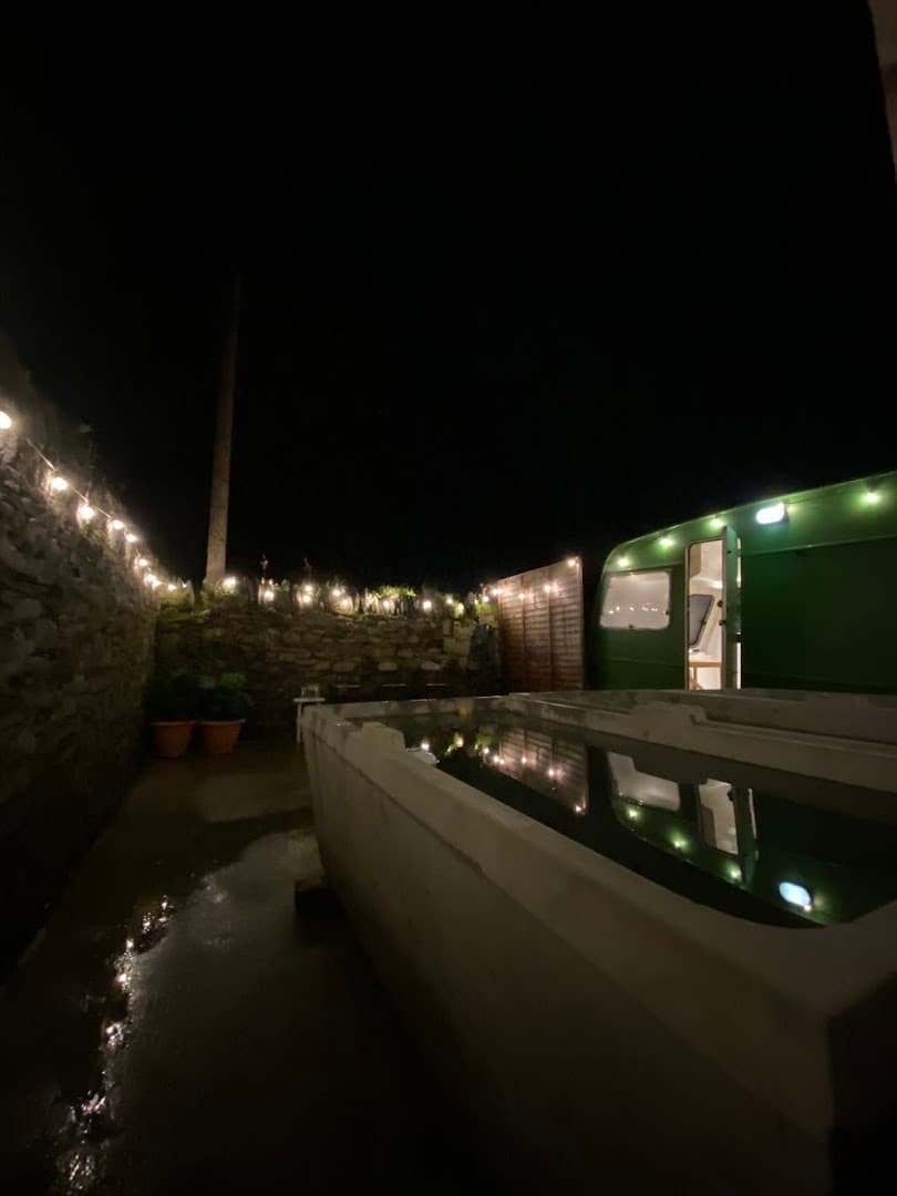 A nighttime outdoor view features a stone wall, string lights, and a green sauna structure next to a tub of water.