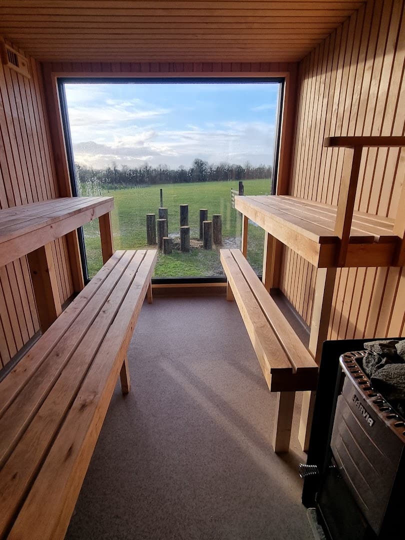 Interior view of a wooden sauna featuring slatted benches, a stove on the right, and a large window framing a grassy field.