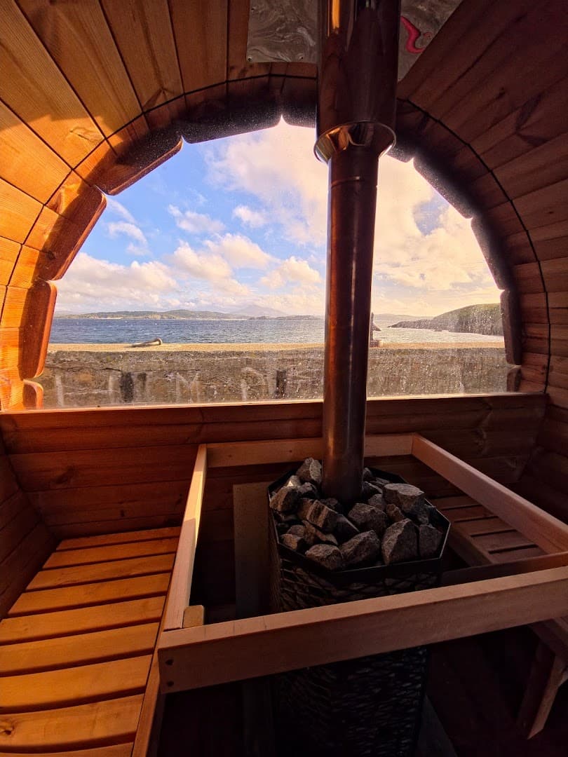 Inside a wooden barrel sauna overlooking the sea, a stove filled with rocks sits beside a glass window.